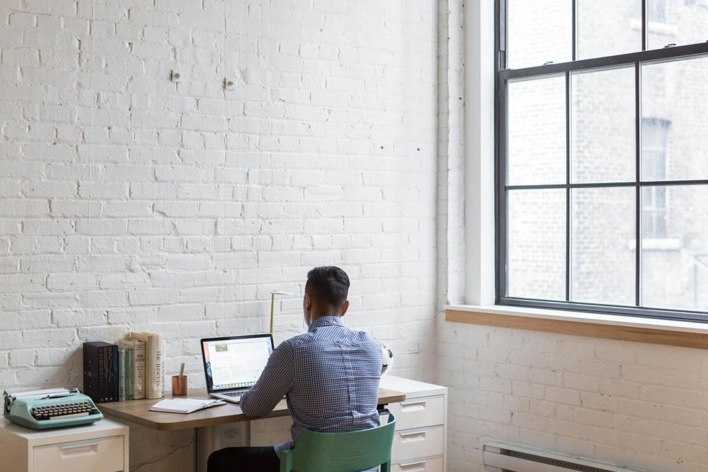 A man sitting at a desk with a laptop and books, facing a large window