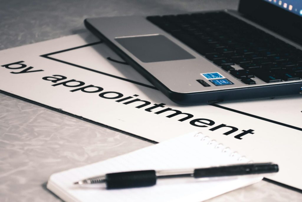 A laptop, a "by appointment" sign, and a notepad with a pen on a desk