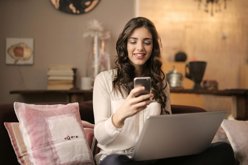 A woman sitting on a couch, smiling while using her phone and laptop