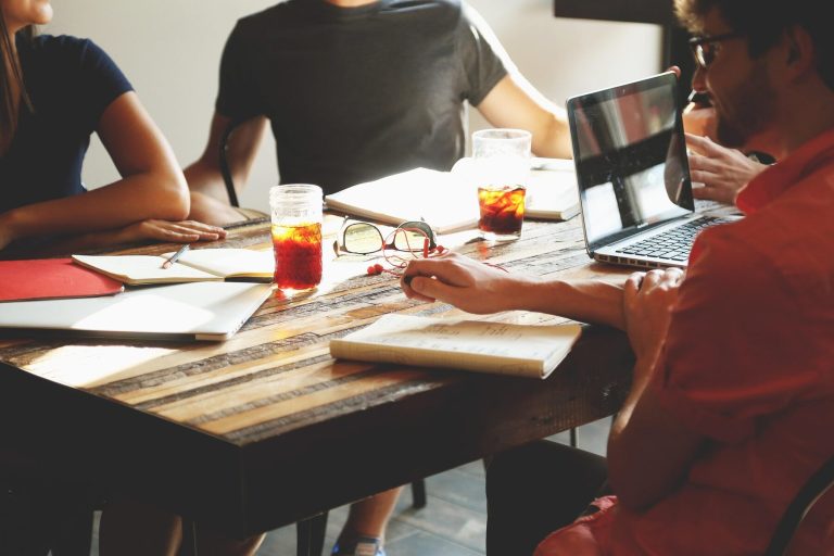 A group of people sitting at a wooden table in a casual meeting setting, with drinks, laptops, and notebooks