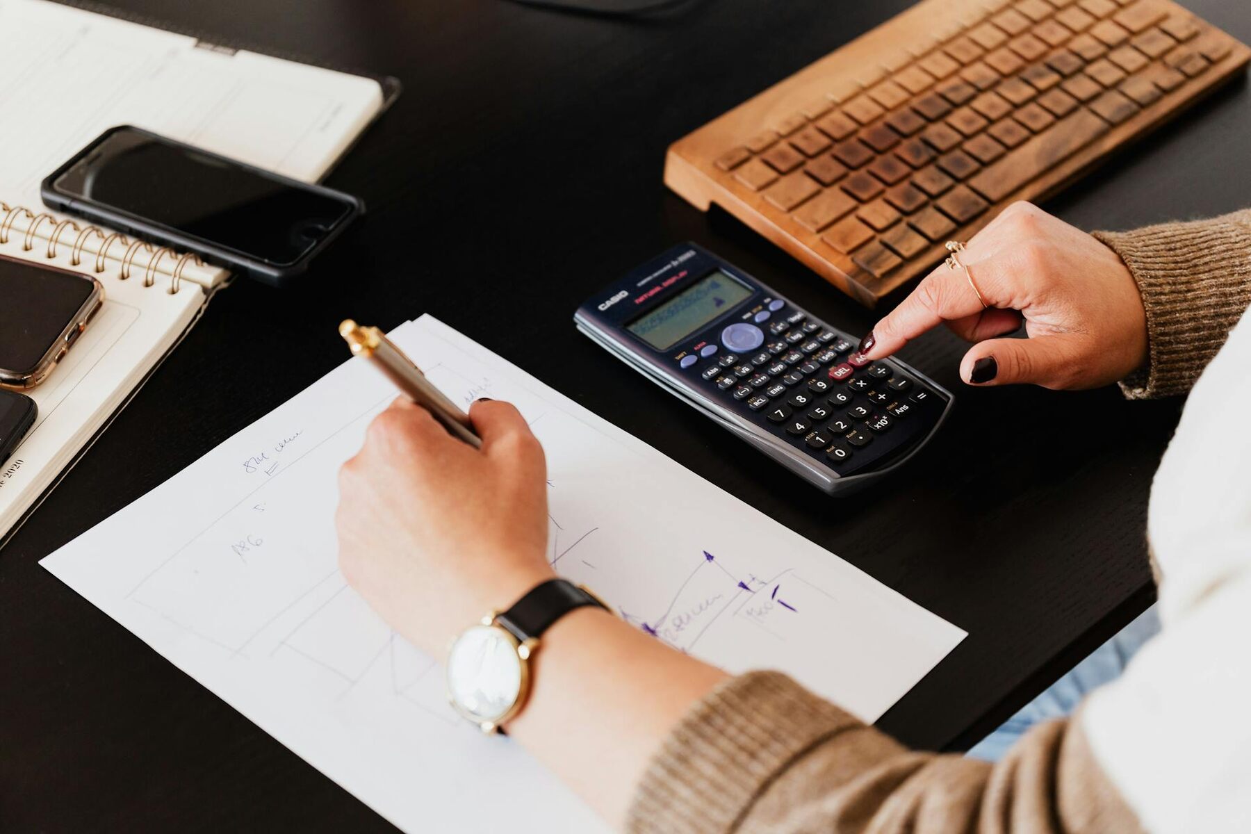 A woman calculates with a calculator while writing notes on paper at a desk